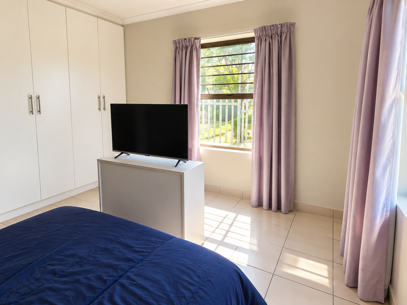 A bright bedroom featuring built-in cupboards, tiled flooring, and large windows with security bars, offering natural light and a clean, functional layout.