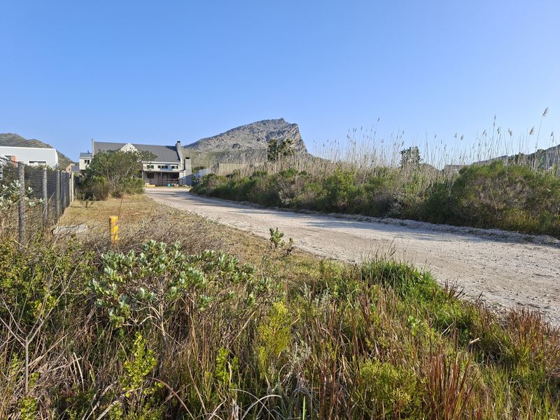 A view of an unpaved access road leading towards a residential property, surrounded by natural vegetation and mountain views.
