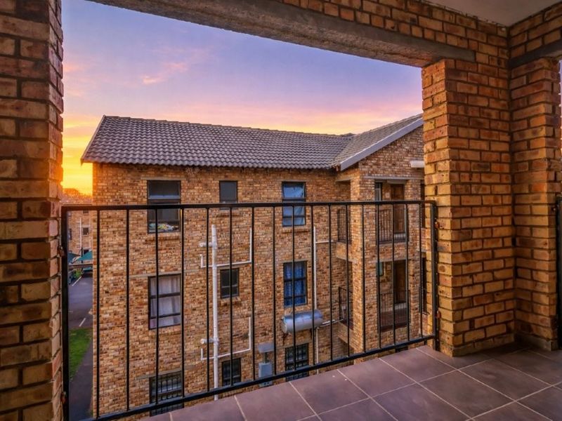 A covered balcony featuring brick pillars, tiled flooring, and a metal balustrade, offering an elevated view of the surrounding apartment complex at sunset.