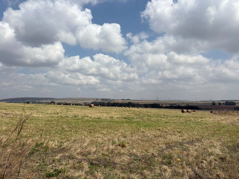 Expansive view of open farmland with rolling fields and hay bales under a bright, cloudy sky.