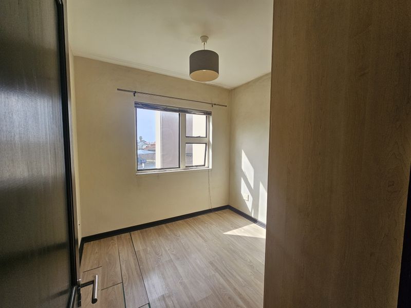 A bright, empty bedroom featuring modern laminate flooring, neutral walls, and a window with a curtain rod, offering a clean canvas for personalization.