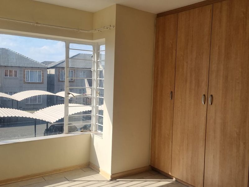 A bright, empty bedroom featuring built-in wooden cupboards, tiled flooring, and large windows with security bars overlooking the complex.