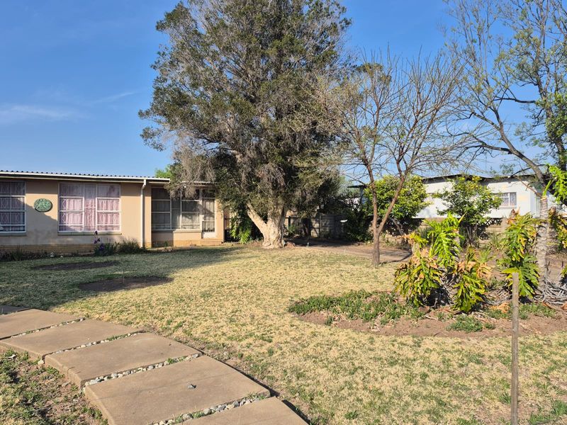 A spacious rear garden featuring mature trees, a concrete pathway, and a view of the property's rear elevation with security-fitted windows.