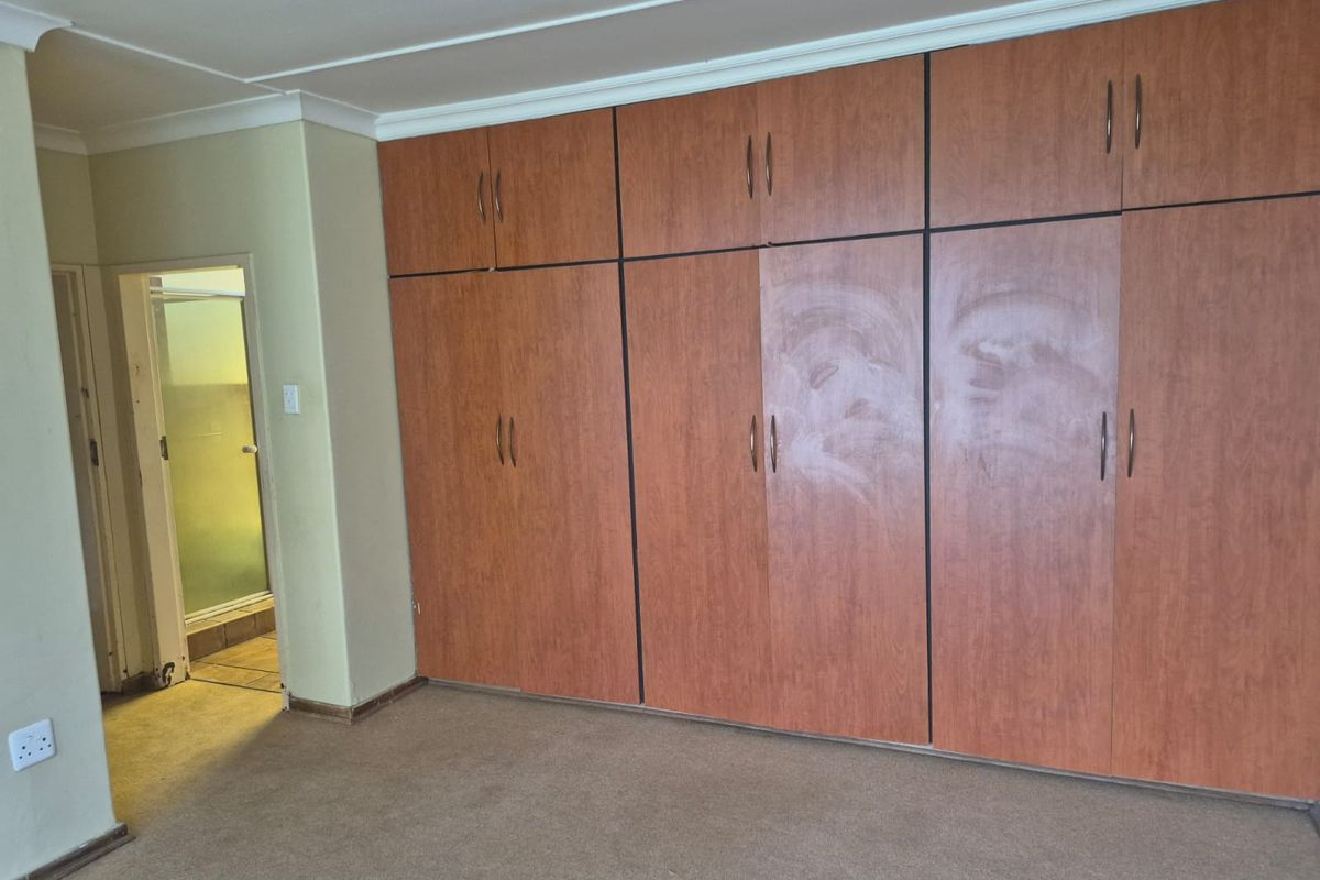 A spacious bedroom featuring extensive built-in wooden cupboards, neutral wall finishes, and carpeted flooring, with an adjacent doorway leading to an en-suite area.