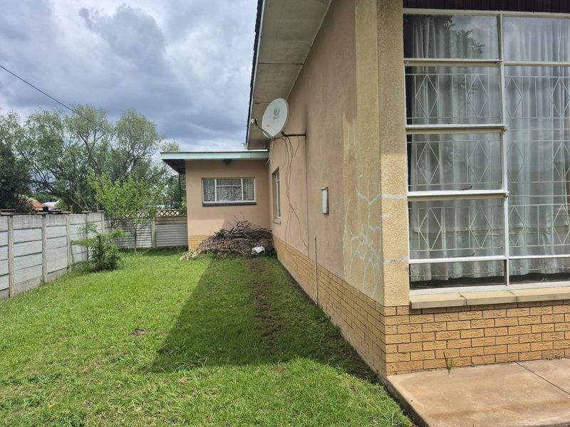 Rear exterior view of the property featuring a lawn, precast boundary wall, and brick plinth with visible plaster cracking and exterior maintenance requirements.