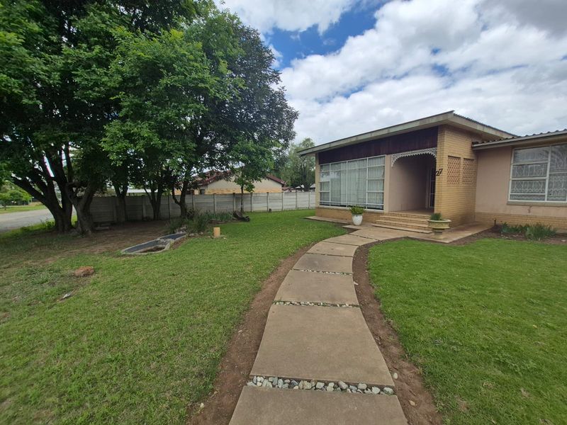 Front view of a brick house featuring a concrete walkway leading to a covered porch, set within a spacious garden with mature trees.