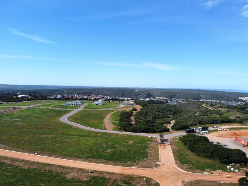 An aerial view of a residential development site featuring paved and dirt roads, surrounding natural vegetation, and distant housing.