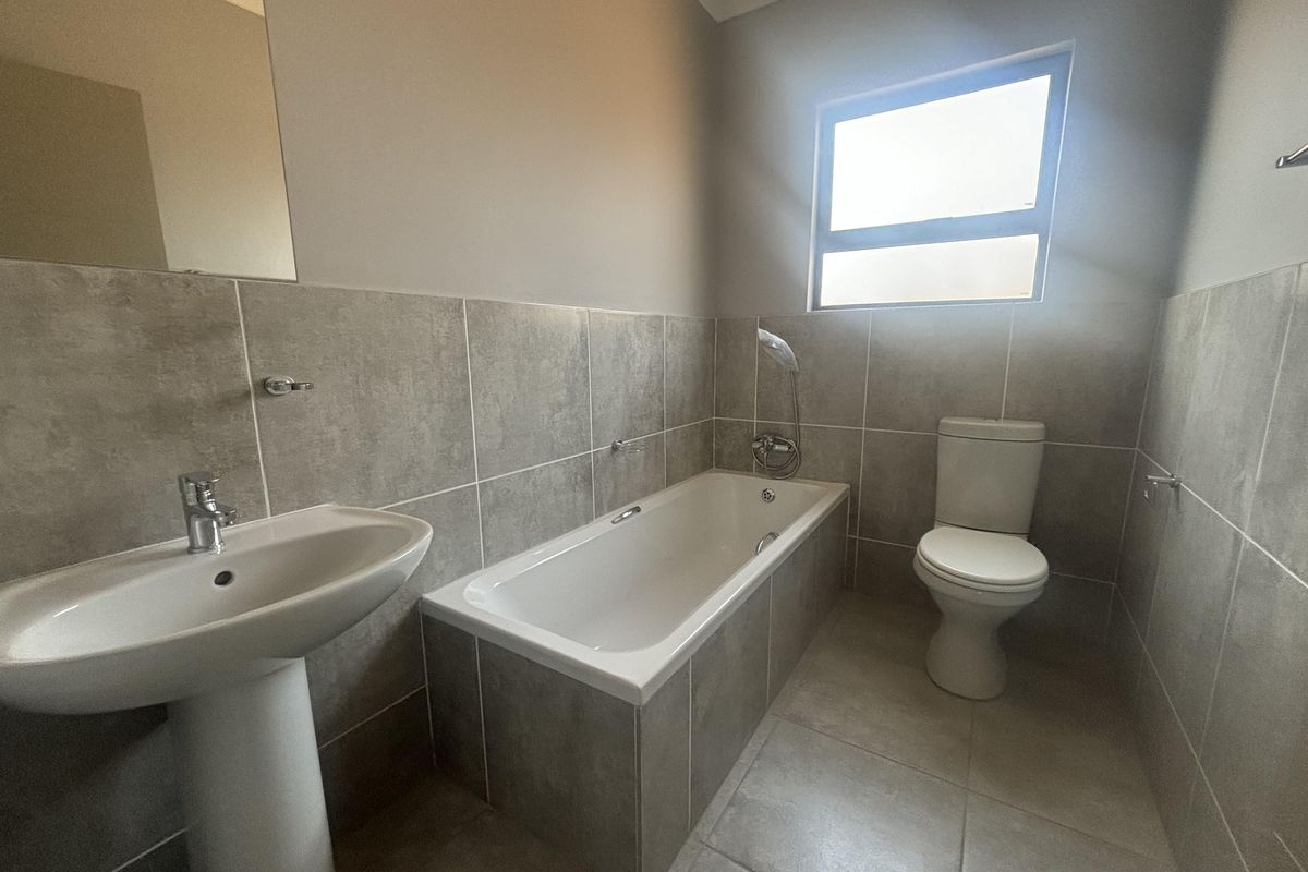 A clean, modern bathroom featuring a pedestal basin, built-in bathtub with handheld shower, and a toilet, finished with neutral grey wall and floor tiles.