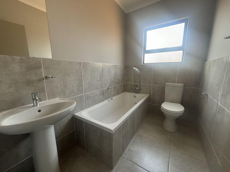 A clean, modern bathroom featuring a pedestal basin, built-in bathtub with handheld shower, and a toilet, finished with neutral grey wall and floor tiles.