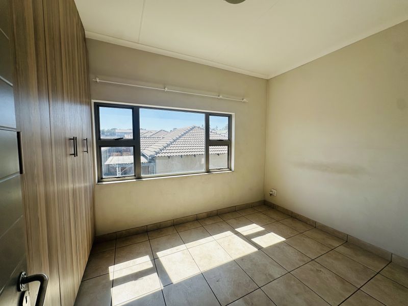A bright, empty bedroom featuring tiled flooring, built-in cupboards, and an aluminium-framed window with a view of the surrounding neighbourhood.