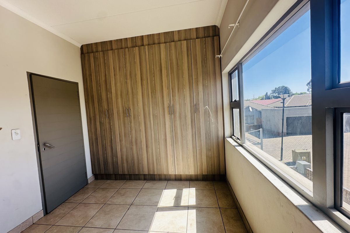 A bright, empty bedroom featuring modern wood-finish built-in cupboards, tiled flooring, and large aluminium-framed windows providing natural light.