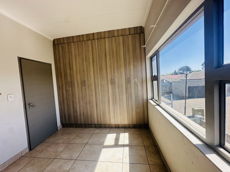 A bright, empty bedroom featuring modern wood-finish built-in cupboards, tiled flooring, and large aluminium-framed windows providing natural light.