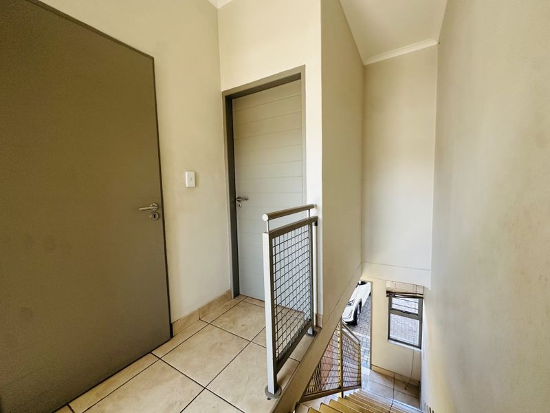A clean, neutral hallway featuring tiled flooring, a metal balustrade, and access to internal rooms, with a staircase leading to the lower level.