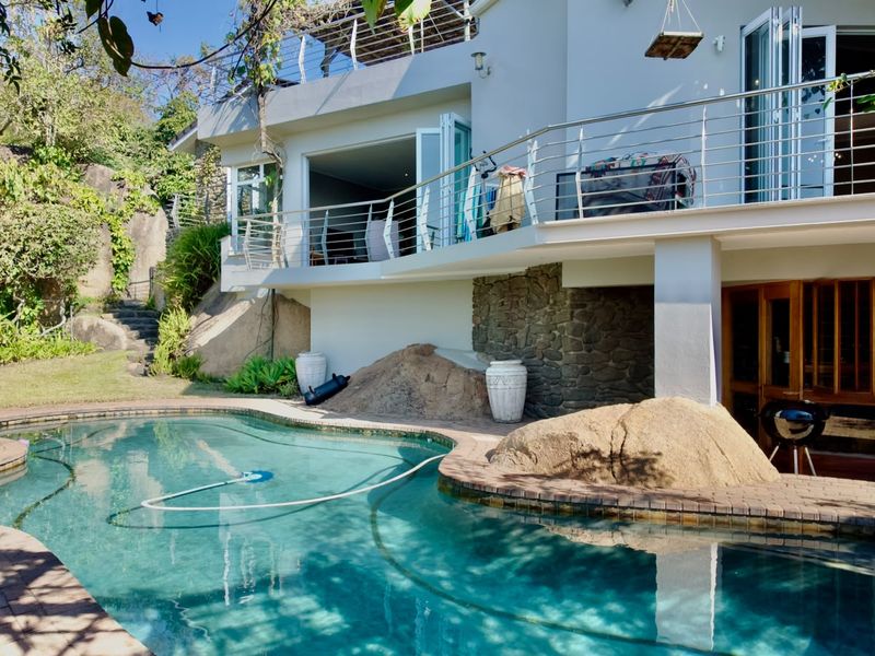 A well-maintained outdoor pool area featuring natural rock landscaping, a paved deck, and a balcony with stainless steel balustrades overlooking the pool.