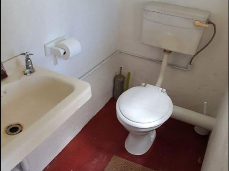 A small, dated bathroom featuring a basic toilet and wall-mounted washbasin with exposed plumbing and worn flooring.