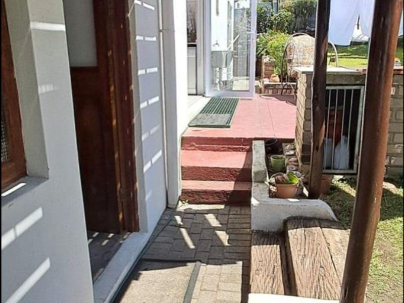A covered outdoor patio area featuring a paved walkway, timber support posts, and steps leading to the rear garden entrance.