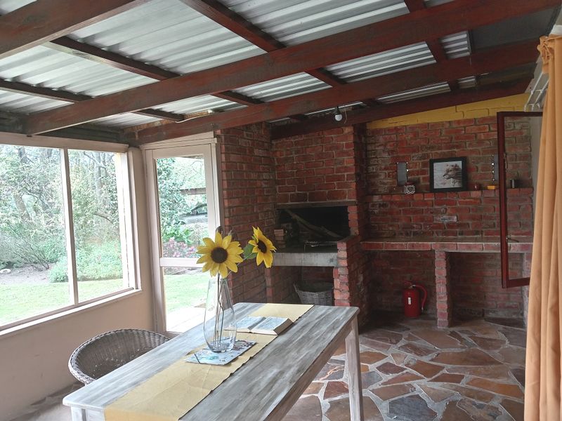 Covered outdoor patio featuring a built-in brick braai, exposed timber beams, and stone flooring, offering a rustic entertainment space overlooking the garden.