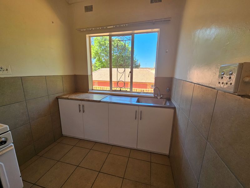 A functional, empty kitchen featuring built-in cabinetry, a stainless steel sink, tiled splashbacks, and tiled flooring with a window providing natural light.