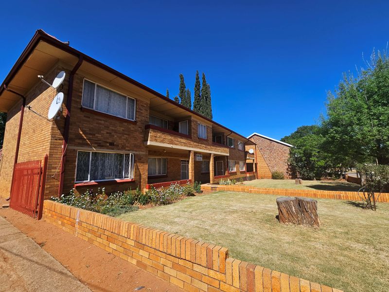 Front exterior view of a multi-story brick apartment building featuring a front garden, brick boundary wall, and paved access area.