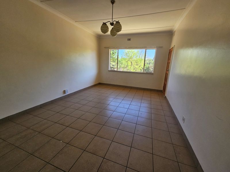 A spacious, empty living room featuring neutral tiled flooring, a large window providing natural light, and a classic ceiling light fixture.