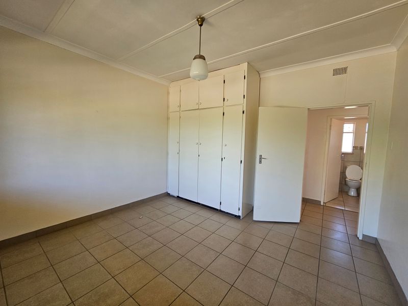 A spacious, empty bedroom featuring neutral tiled flooring, built-in cupboards, and a view into the adjacent bathroom.