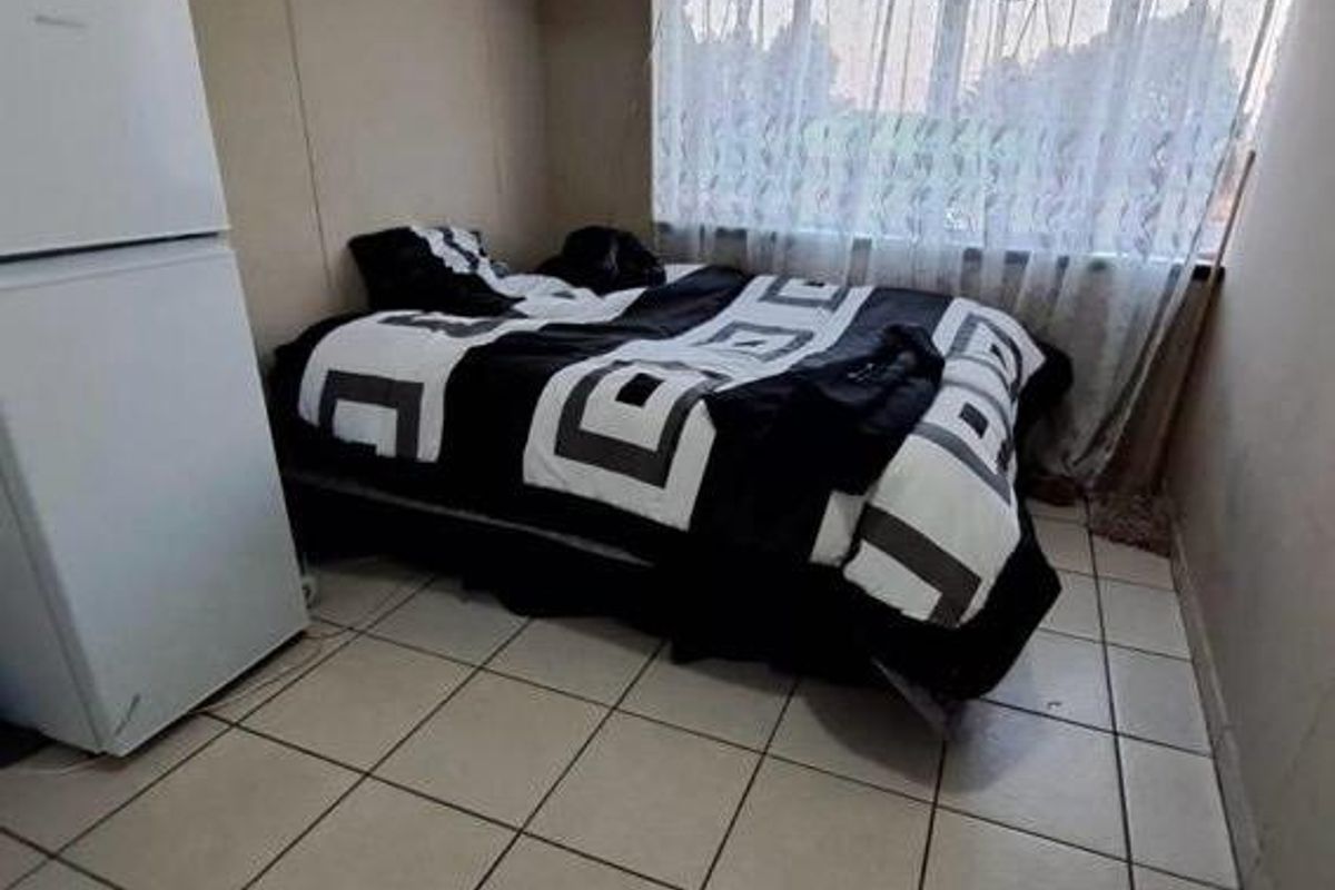 A compact bedroom featuring tiled flooring and a large window with security bars, requiring cosmetic updates to improve the overall aesthetic.