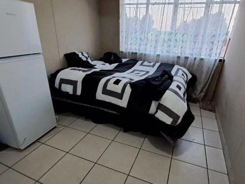 A compact bedroom featuring tiled flooring and a large window with security bars, requiring cosmetic updates to improve the overall aesthetic.