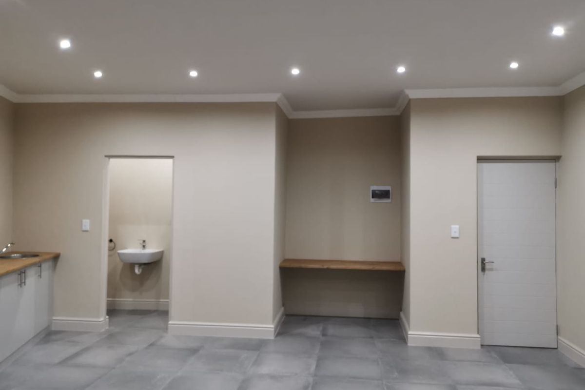 A modern, neutral-toned open plan space featuring tiled floors, recessed lighting, a small kitchenette area, and a separate washroom with a basin.
