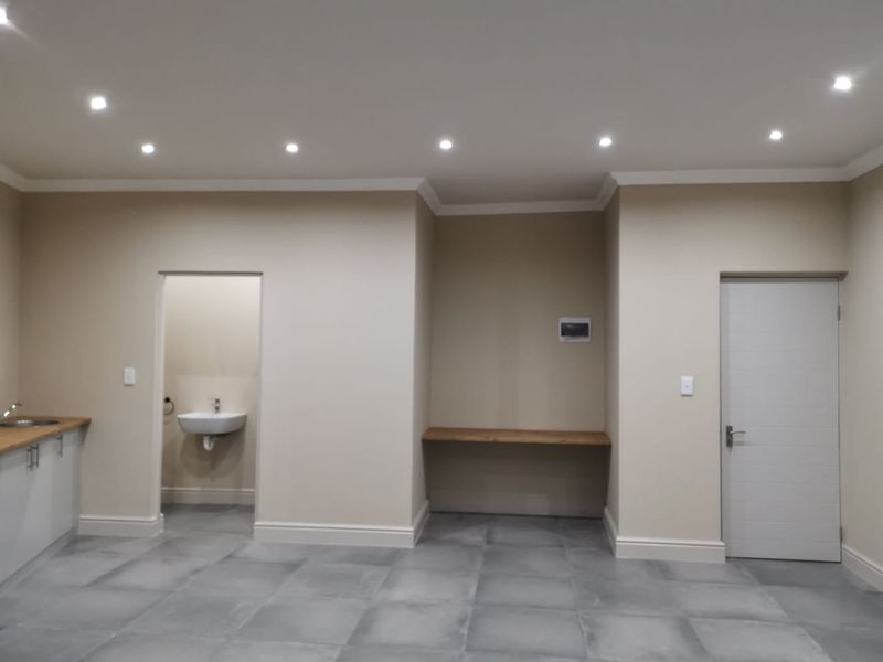 A modern, neutral-toned open plan space featuring tiled floors, recessed lighting, a small kitchenette area, and a separate washroom with a basin.