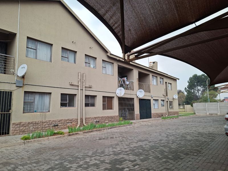 Exterior view of a multi-story flat complex featuring paved driveway, shade sail parking, and balcony access.