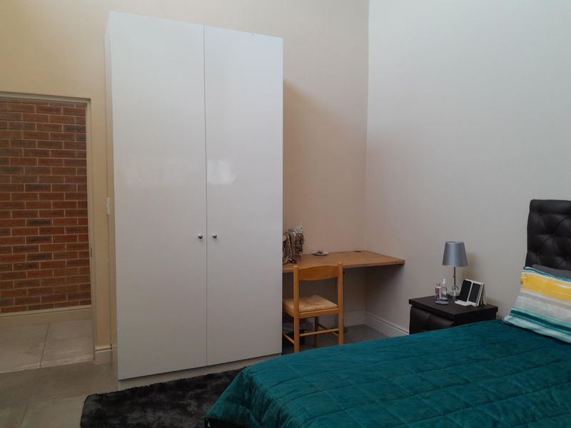 A bedroom featuring a white built-in wardrobe and a built-in desk area, with access to an adjacent room with exposed brickwork.