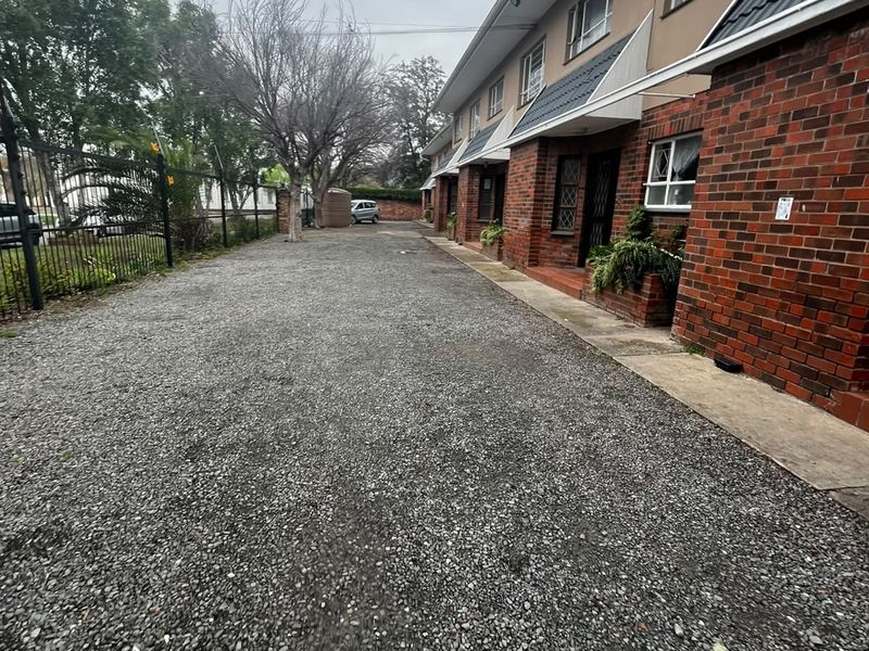 Exterior view of an apartment complex featuring a brick facade, tiled roof, and a gravel driveway with perimeter security fencing.