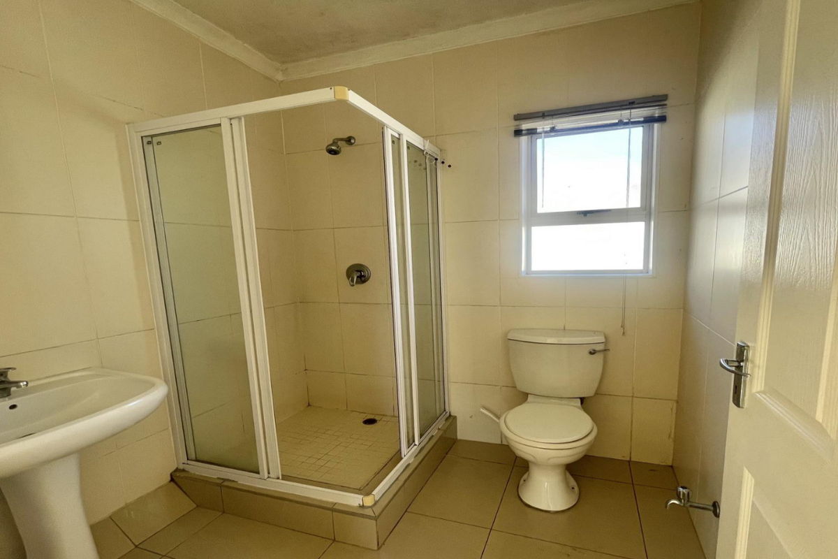 A functional bathroom featuring a pedestal basin, a corner shower enclosure, and a toilet, finished with neutral wall and floor tiles.