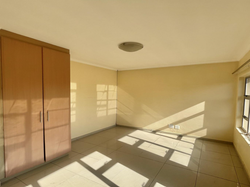 A bright, empty bedroom featuring neutral tiled flooring, a built-in wooden wardrobe, and large windows with curtain rails.