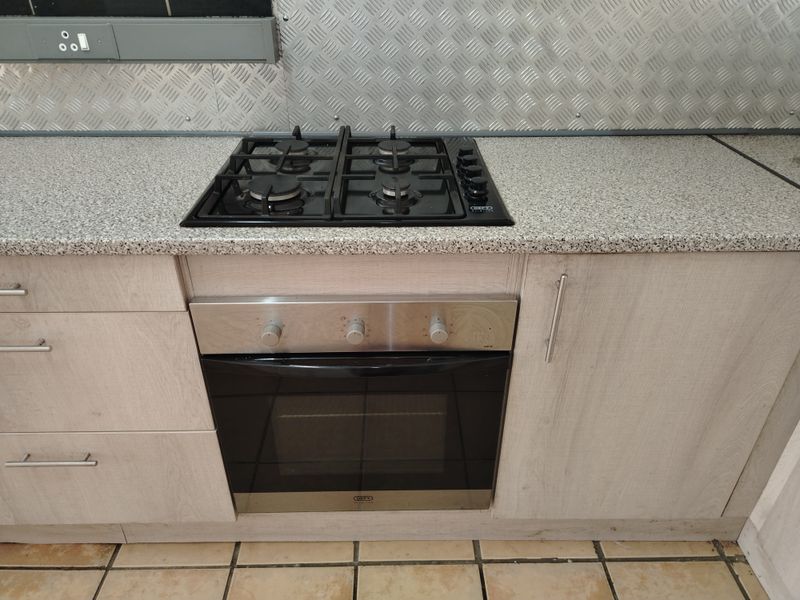 A functional kitchen area featuring a built-in gas hob, stainless steel oven, light wood-look cabinetry, and a durable metal splashback.