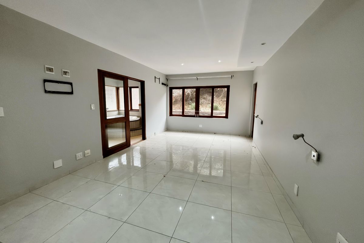 A spacious, empty bedroom featuring neutral tiled flooring, recessed lighting, and wooden window frames, with an adjoining room visible through a sliding door.