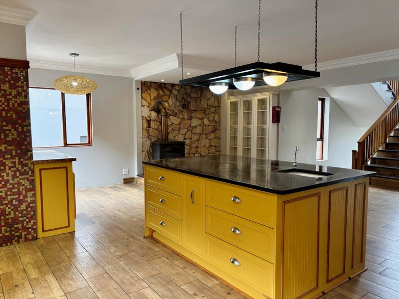 Spacious open-plan living area featuring a vibrant yellow kitchen island, stone fireplace with wood-burning stove, and a timber staircase.