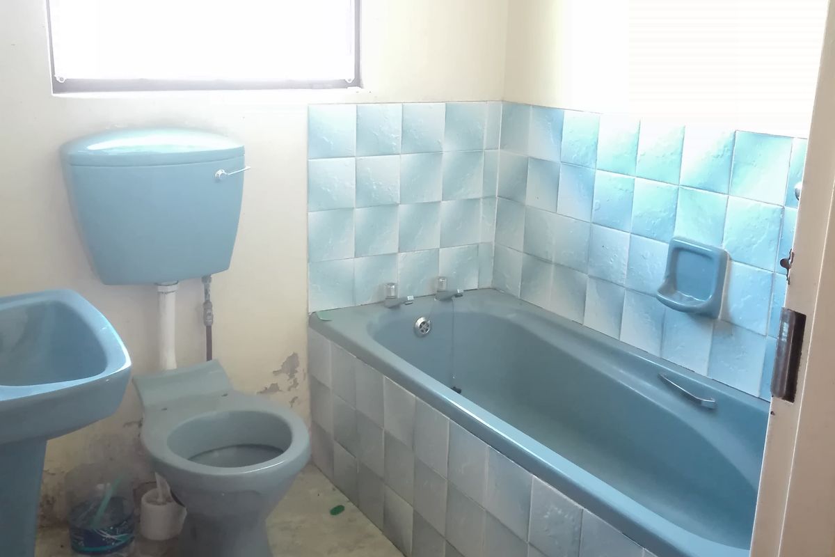 A dated bathroom featuring blue sanitaryware, tiled bathtub surround, and a window with security bars, requiring modernization and cosmetic updates.