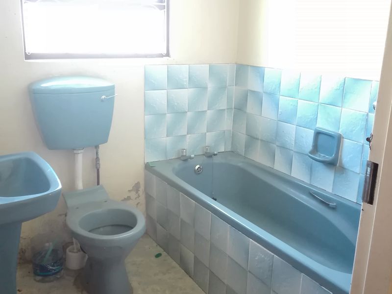 A dated bathroom featuring blue sanitaryware, tiled bathtub surround, and a window with security bars, requiring modernization and cosmetic updates.