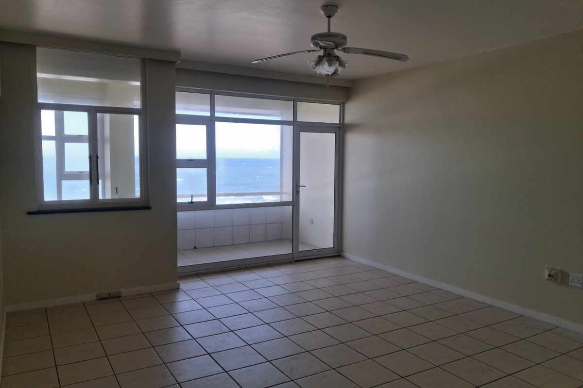 Spacious living area featuring tiled flooring, a ceiling fan, and large windows providing direct access to a balcony with ocean views.