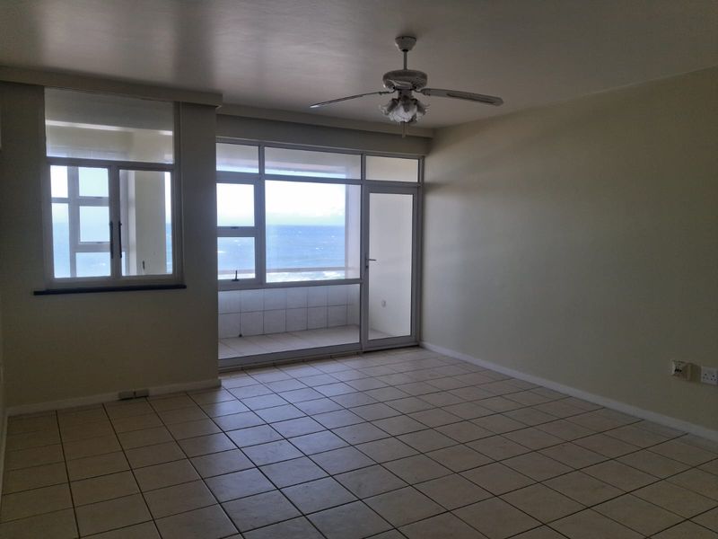 Spacious living area featuring tiled flooring, a ceiling fan, and large windows providing direct access to a balcony with ocean views.