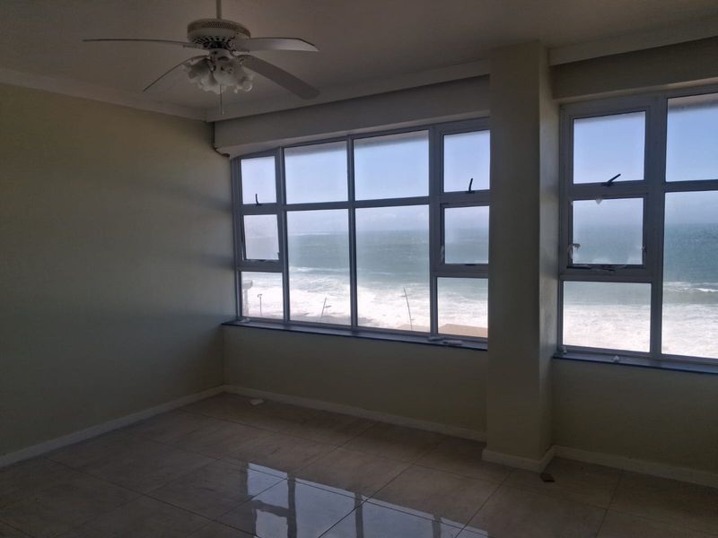 An empty, light-filled living area featuring tiled floors, a ceiling fan, and expansive windows offering direct ocean views.