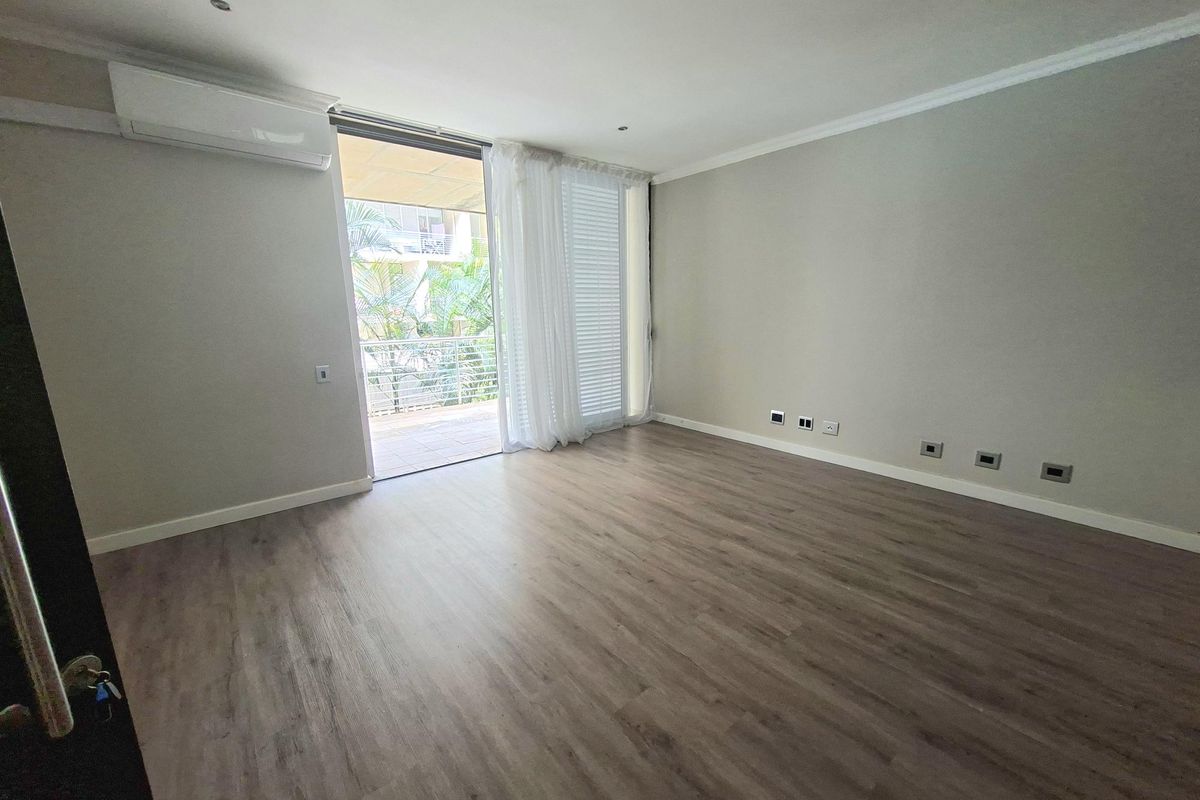 A bright, empty living room featuring modern laminate flooring, a split-system air conditioner, and direct access to a private balcony through sliding glass doors.