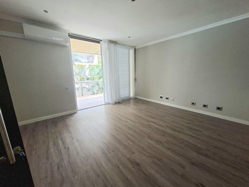 A bright, empty living room featuring modern laminate flooring, a split-system air conditioner, and direct access to a private balcony through sliding glass doors.