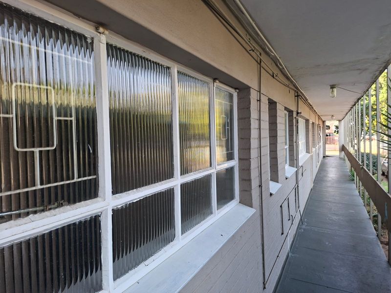 An exterior apartment walkway featuring painted brick walls, ribbed glass windows with security bars, and exposed electrical conduit.