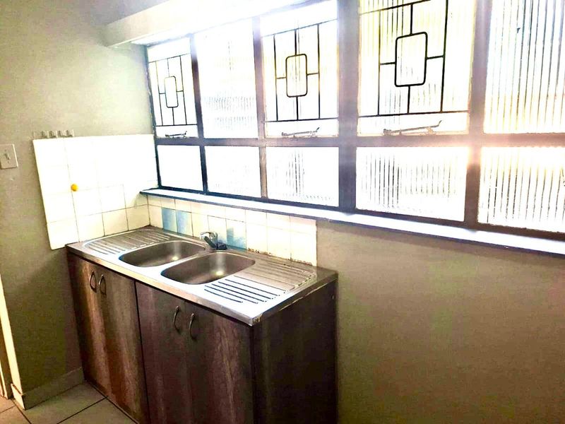 A compact kitchen area featuring a stainless steel double sink, built-in cabinetry, and tiled splashback, illuminated by large windows with security bars.