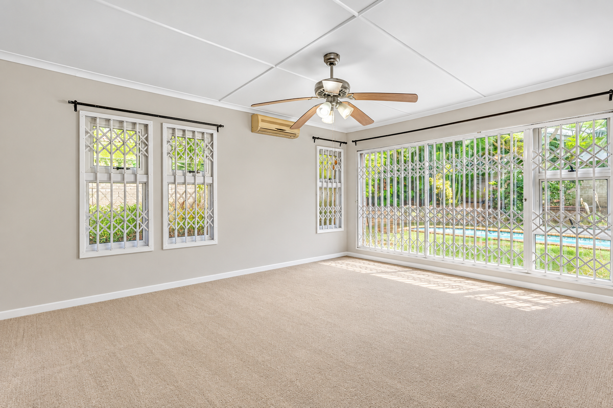 Spacious, light-filled living room featuring neutral carpeting, a ceiling fan, split-system air conditioning, and large windows with security gates overlooking the garden.