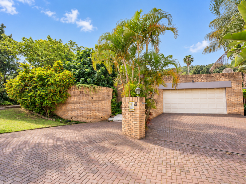Front view of the property featuring a paved driveway, double garage, and established brick facade surrounded by lush garden landscaping.