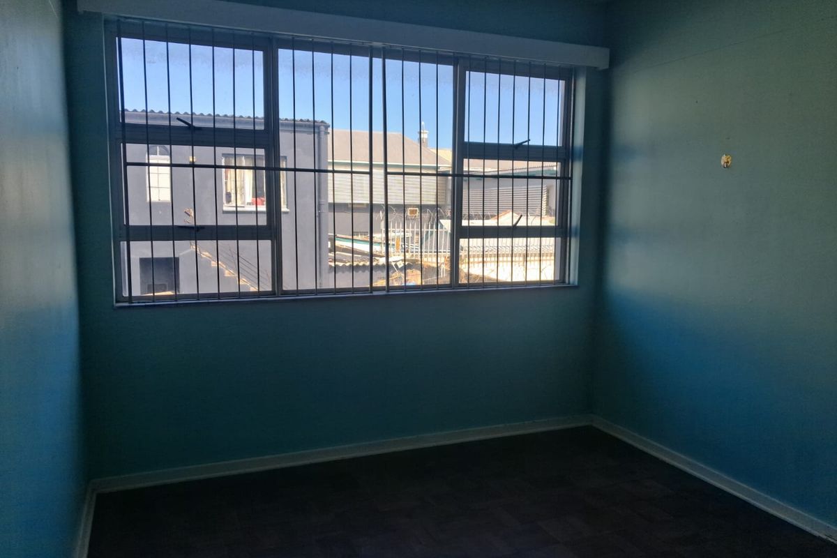 An empty bedroom featuring parquet flooring and large windows fitted with security bars, offering a view of the neighboring building.