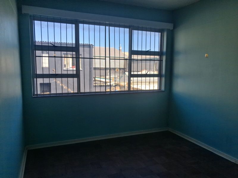 An empty bedroom featuring parquet flooring and large windows fitted with security bars, offering a view of the neighboring building.